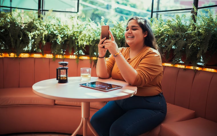 A smiling person sitting at a table in a coffee shop and looking at a phone in their hand